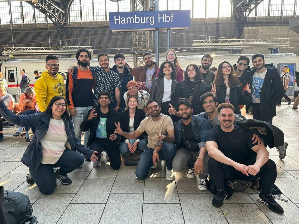 Eine multikulturelle Gruppe von jungen Studierenden der Hochschule Niederrhein und Mitarbeiterinnen des International Office lachen in die Kamera am Hamburger Hauptbahnhof.