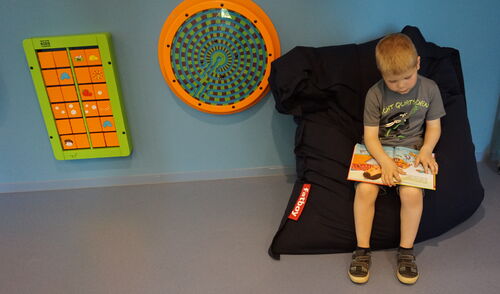 Kind sitzt auf einem Sitzsack und liest ein Buch. Neben dem Kind hängen Spielgeräte an der Wand.