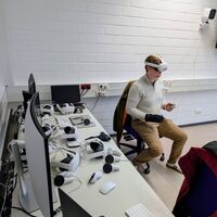 Eine Person mit VR Brille, Controller und einem speziellen Handschuh sitzt auf einem Bürostuhl. Auf dem Schreibtisch daneben sind Computer und weitere VR-Brillen zu sehen.