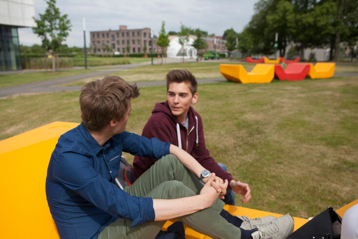 Zwei Studenten sitzen auf den buten Sitzgelegenheiten im Außengelände des Campus Krefeld Süd und sind ins Gespräch vertieft.