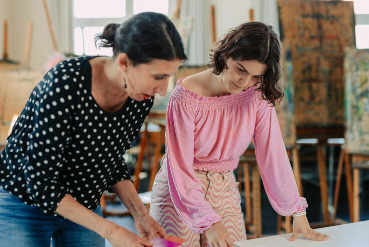 A student in a pink blouse bends over design drafts with her lecturer. In the background you can see colorful canvases on easels.