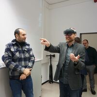 2 Personen in einem Raum, eine Person hat eine VR Brille an und zeigt auf etwas, was nur in der VR sichtbar ist.