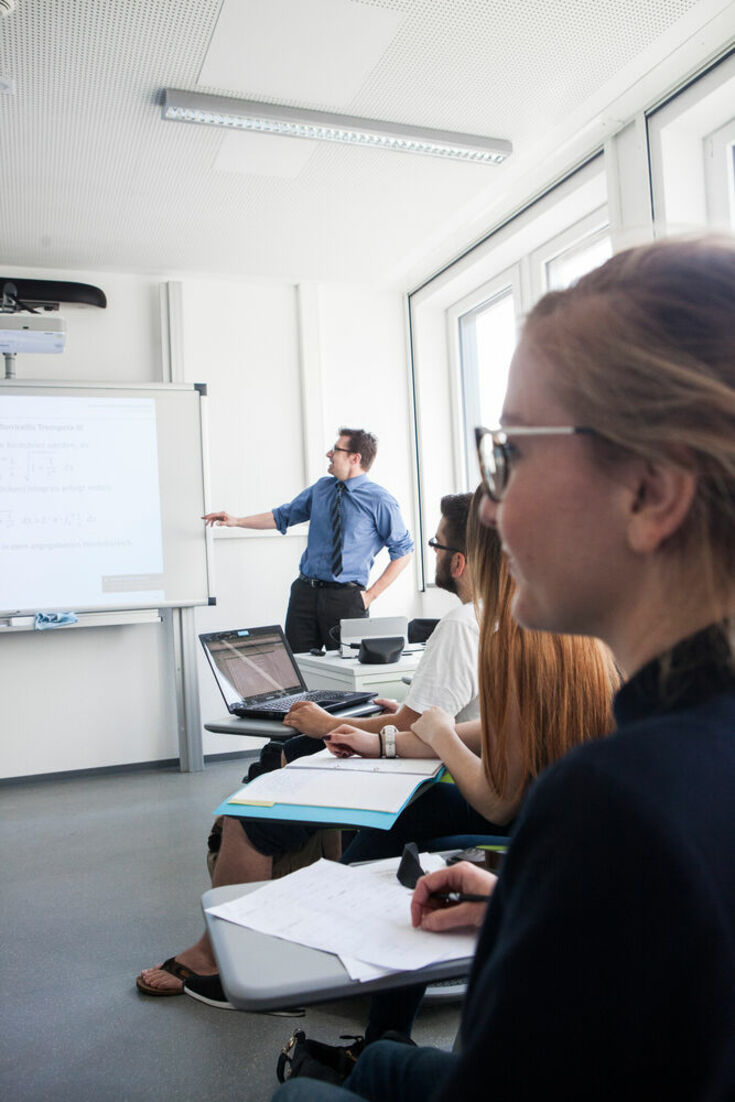 Students are sitting in the seminar room; a university teacher / lecturer stands in front of the whiteboard and points to it. 