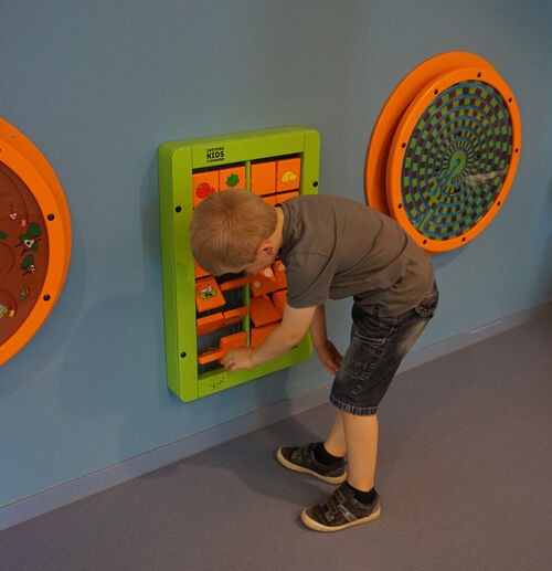 Ein Kind spielt mit einem Spielgerät, welches an der Wand montiert ist. Links und rechts neben dem Kind hängen weitere Spielgeräte an der Wand.