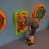 Ein Kind spielt mit einem Spielgerät, welches an der Wand montiert ist. Links und rechts neben dem Kind hängen weitere Spielgeräte an der Wand.