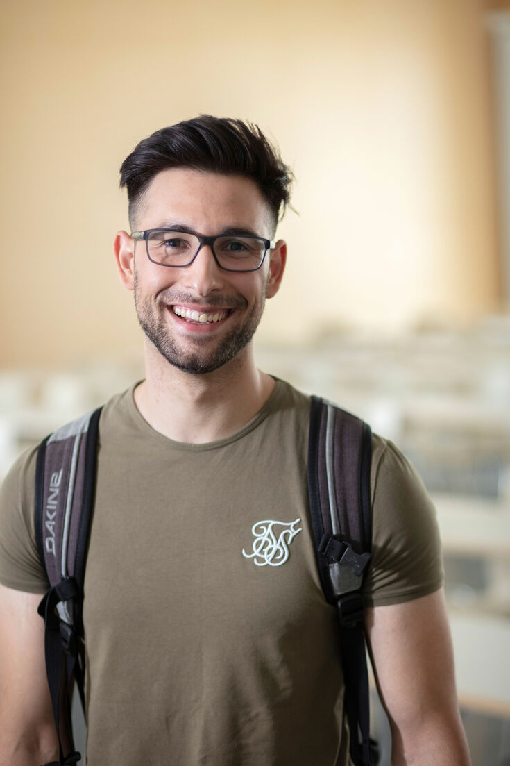 Ein Student mit moderner dunkler Hornbrille und Rucksack lächelt in die Kamera.