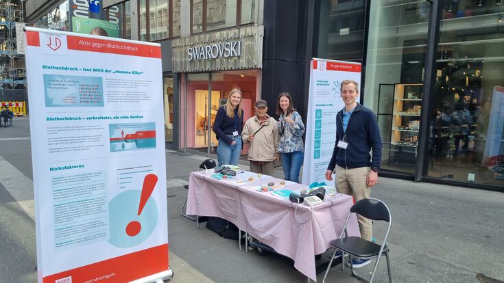 Lukas Streese und 3 weitere Personen am Infostand. Man sieht die Rollups und den Tisch mit Infomaterial sowie mehrere Blutdruckmessgeräte